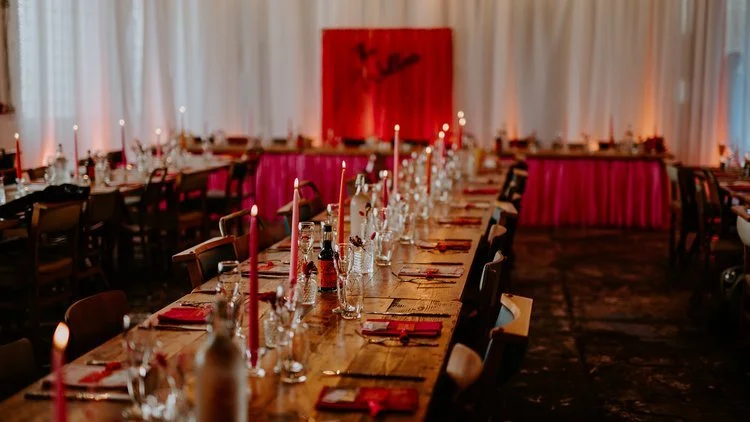 A warm and atmospheric wedding reception setup by The Village Green Events, Sheffield. The industrial-style venue features long wooden banquet tables adorned with pink taper candles, glassware, and personalised place settings. A striking red backdrop
