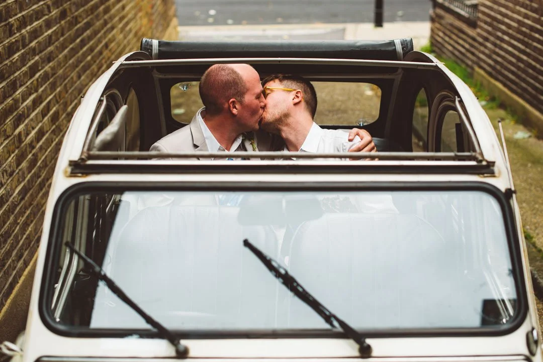 A tender and characterful moment captured by Barbara K Photography, showing two grooms sharing a kiss inside a vintage car with the roof open. Photographed from above, the image frames their embrace through the top of the classic vehicle, creating a 