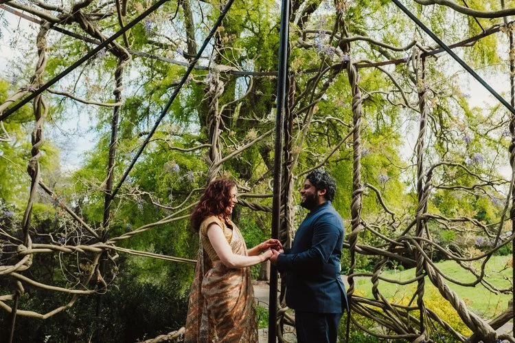A couple stand facing one another beneath an intricate canopy of twisted vines and wisteria, sharing a tender moment during their outdoor celebration. The bride’s richly patterned gown and the groom’s smart suit contrast beautifully with the lush gre