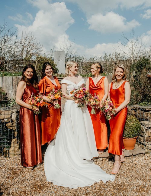 Joyful bridal party holding bright seasonal bouquets by The Art Bunch featuring colourful roses, ranunculus, tulips and fresh garden foliage. With bridesmaids in vibrant orange satin dresses beside the bride in a classic white gown, this lively moder