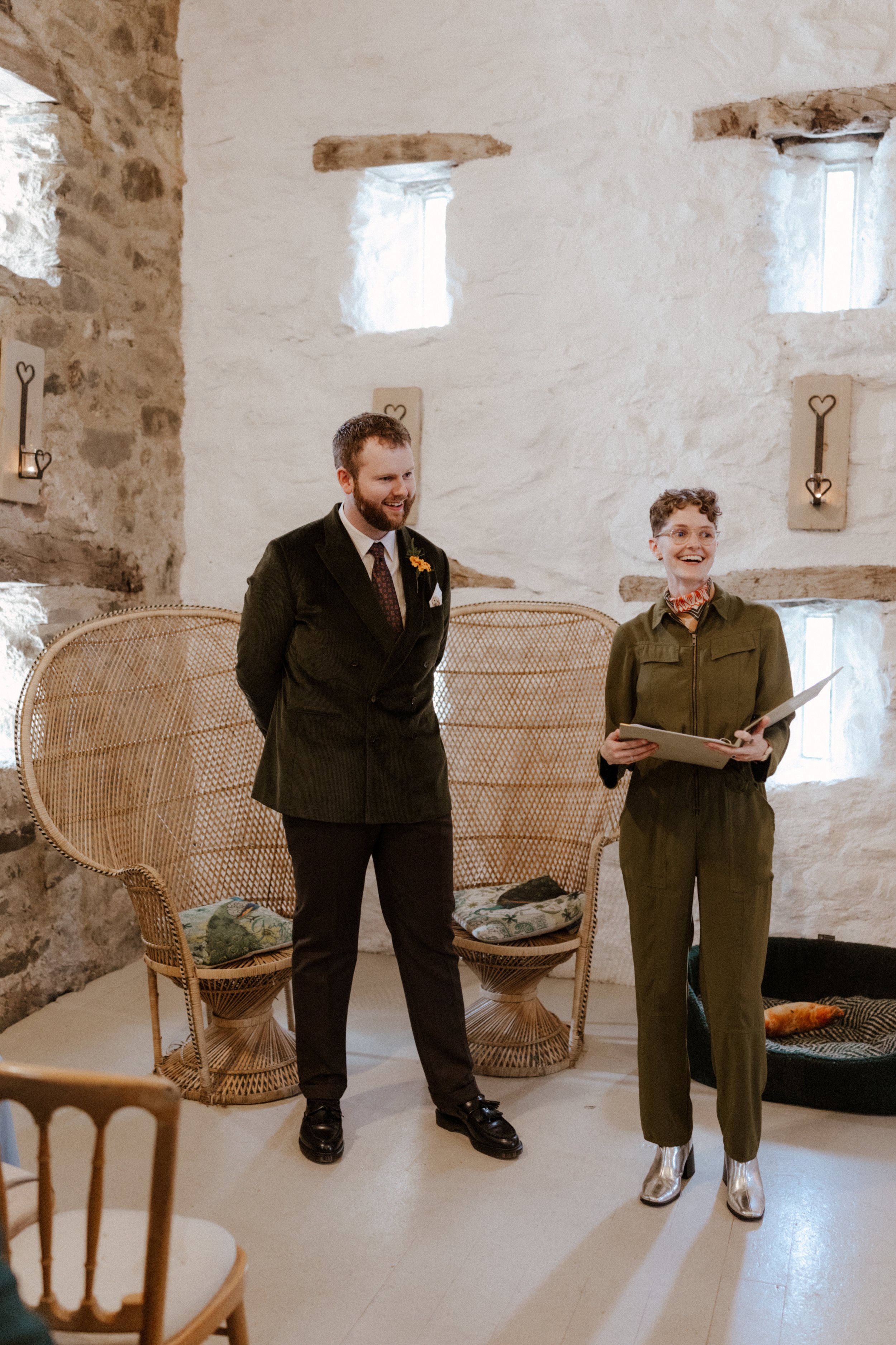A relaxed rustic wedding ceremony in a stone barn featuring a smiling Chloe Green wedding celebrant standing beside the groom before the arrival of the bride. The scene is full of warmth and natural charm, with wicker chairs, soft lighting and earthy