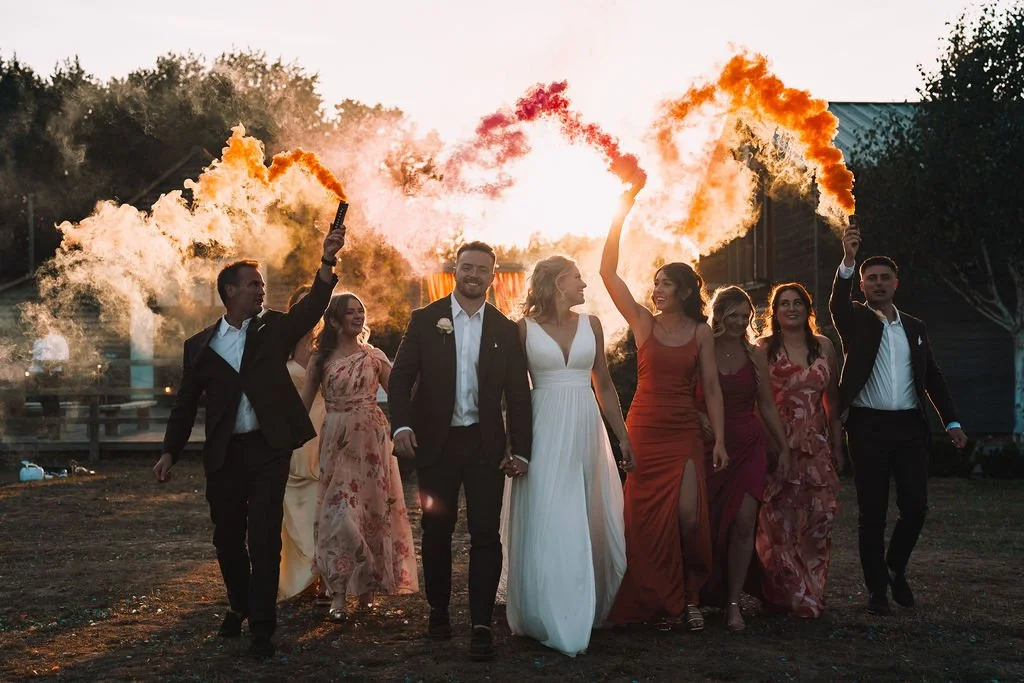 A wedding party walks together through warm evening light as the couple leads the group hand in hand, surrounded by friends in coordinated suits and dresses who hold aloft orange and red smoke flares that billow dramatically overhead while the sun se