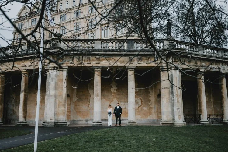 A grand and atmospheric wedding portrait captured by Matt Fox Photography, showing a couple standing together beneath the imposing columns of a historic stone building. The sweeping architecture, weathered textures and curved façade create a dramatic