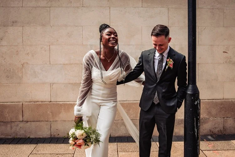 A fun and joy filled wedding moment captured by Natalia Case Photography, showing the couple sharing an unrestrained laugh as they stand together against a warm stone backdrop. The bride’s chic white jumpsuit and sheer sleeves bring a modern, fashion