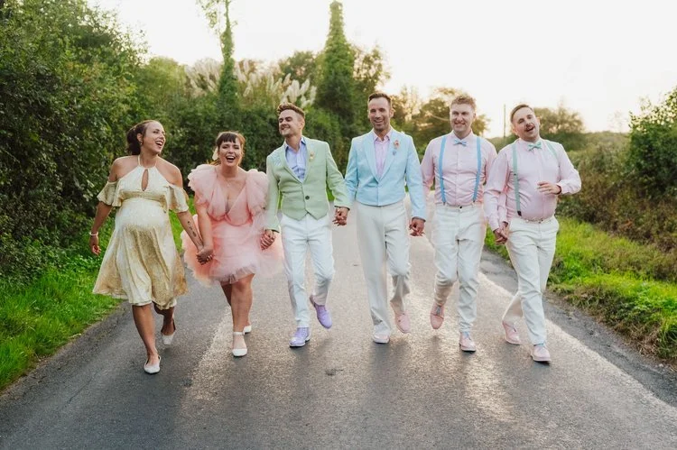 A fun and colourful wedding party walks hand-in-hand down a quiet countryside road, dressed in coordinated pastel outfits. The group laughs together as the golden evening light creates a warm, romantic atmosphere. Captured by Sidey Clarke Photography
