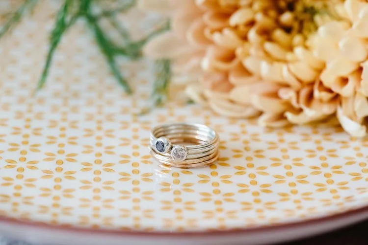 Set of handcrafted silver stacking rings with gemstones by Nikki Stark Jewellery, displayed on a patterned plate beside soft florals. Ethically made in the UK, these elegant rings are perfect for brides seeking bespoke wedding jewellery.