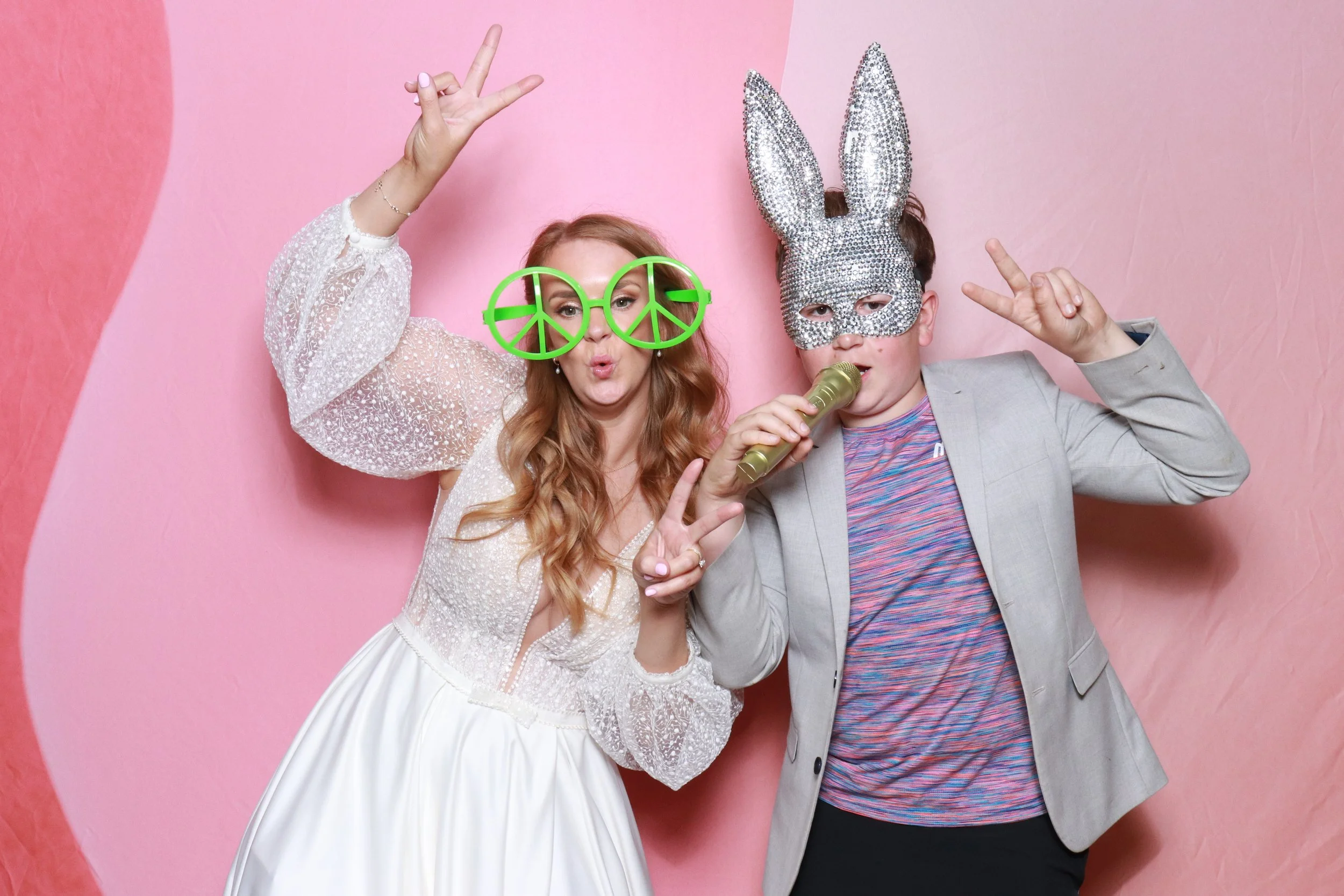 Guests strike a pose in front of Viva La Booth’s vibrant pink backdrop, adding playful props like oversized glasses and glittering masks for a fun photo moment. Perfect for weddings, birthdays, and events, this photobooth creates bright, high-quality