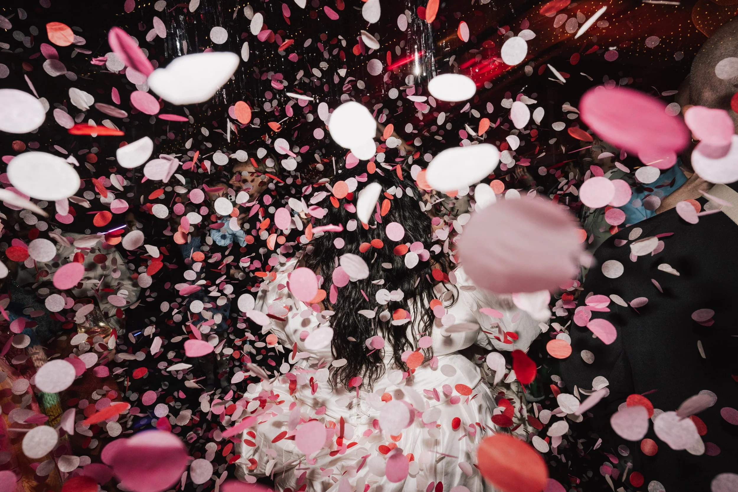 A shower of pink, red, and white confetti fills the frame as a bride celebrates on the dance floor, her flowing hair and satin dress just visible through the swirl of colour. The dramatic lighting and movement create an electric, party-fuelled atmosp