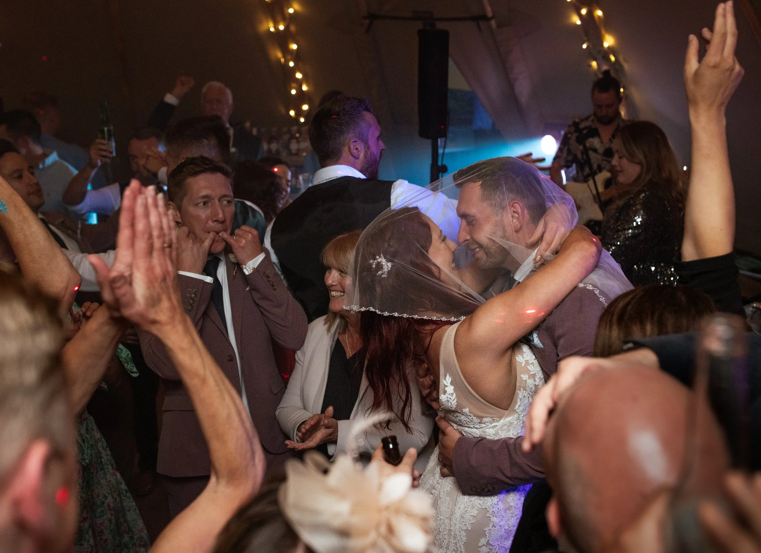 A vibrant, joy-filled wedding party moment captured by Laura Wilson Photography, showing the newly married couple sharing an embrace and smiling beneath the bride’s veil in the middle of a packed dance floor. Surrounded by cheering guests, raised han