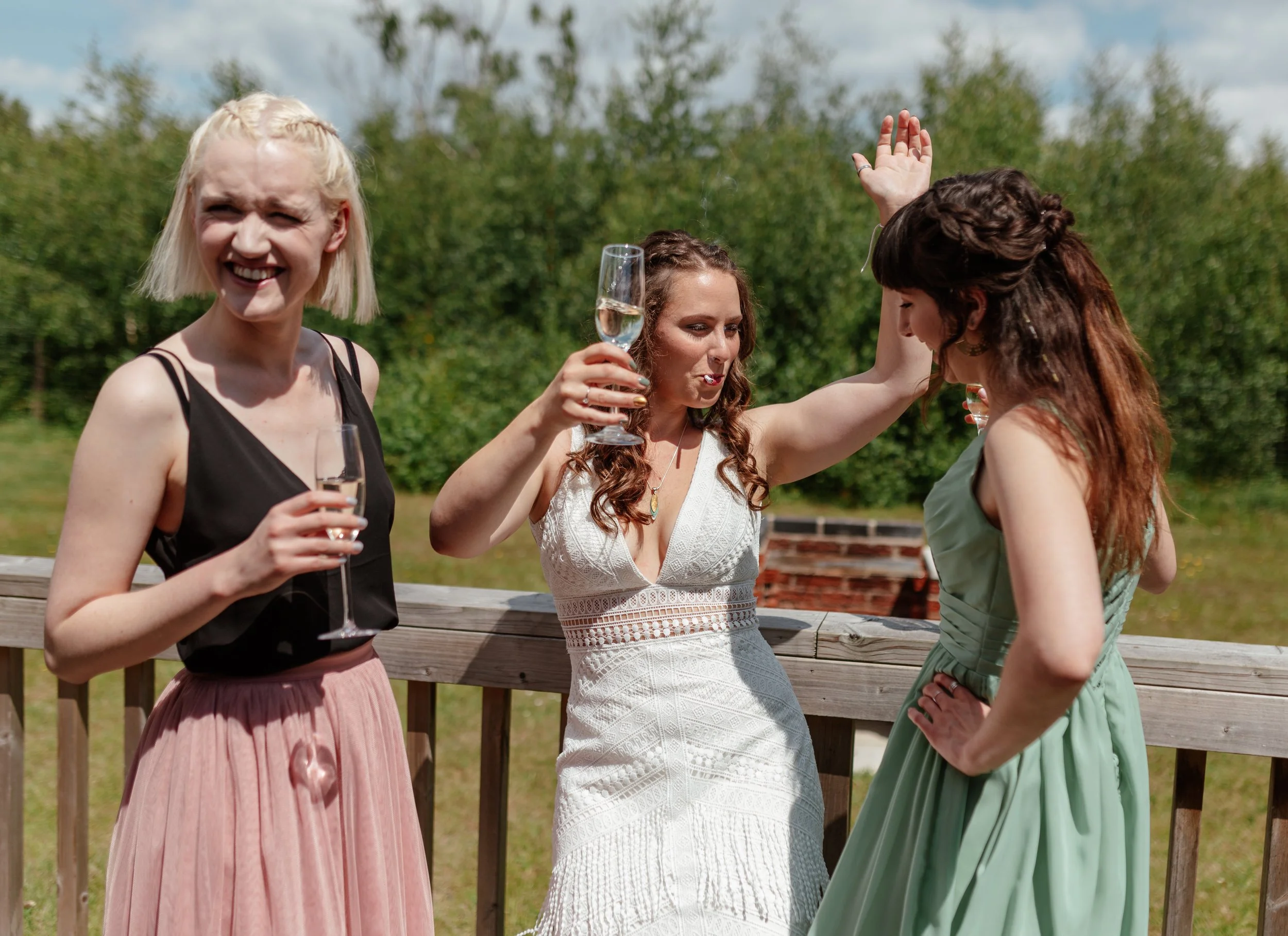 A fun and relaxed wedding moment captured by Laura Wilson Photography, showing the bride and her friends enjoying drinks together in bright sunshine. The bride stands on a wooden deck in a lace dress, lifting her champagne glass and laughing with pla