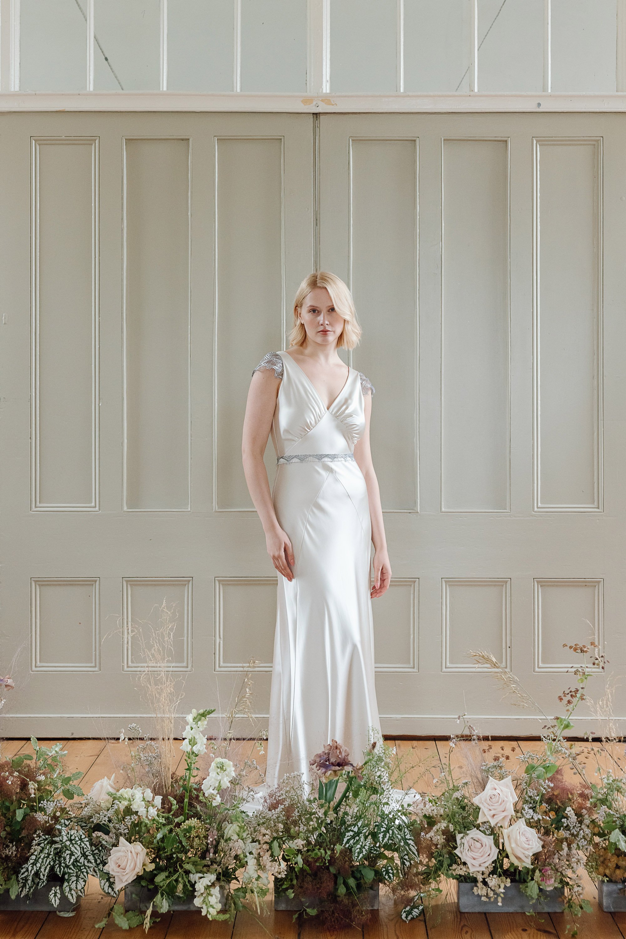 The model stands among soft, garden inspired floral arrangements wearing a bias cut silk gown in a pale, luminous shade, its gentle drape enhanced by a wrapped bodice with a deep V neckline and delicate silver embellished cap sleeves.