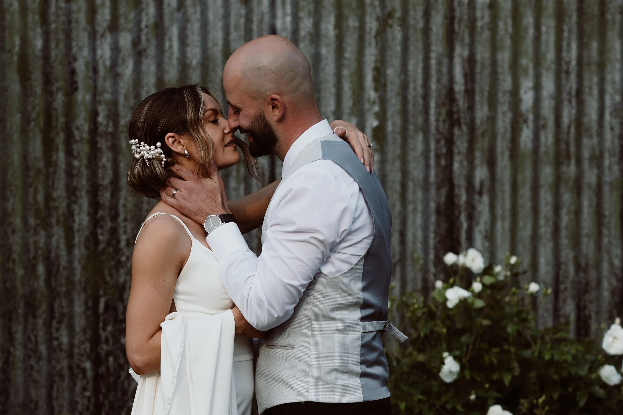 A tender and emotional portrait captured by Joy Louise Photography, showing a couple in a quiet moment of closeness as they lean in for a soft kiss. The groom gently holds the bride’s face while she rests her hands on his shoulders, their expressions