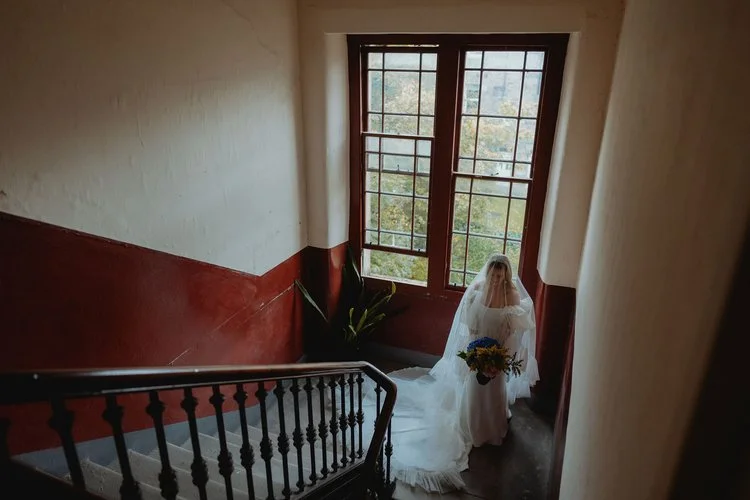 A bride stands at the foot of an old stairwell, framed by tall vintage windows and soft natural light, beautifully captured by F.D Young Photography. Her flowing veil and bouquet contrast with the textured, characterful interior, creating a moody, ci