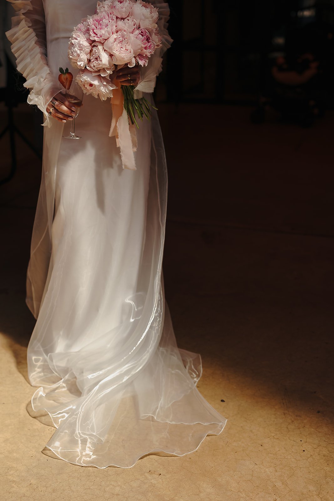 A close view shows the lower half of a white dress made from smooth, opaque fabric layered with a sheer overlay that falls into a soft train on the floor. The hem catches the light, outlining the translucent edges. At waist level, a hand holds a roun