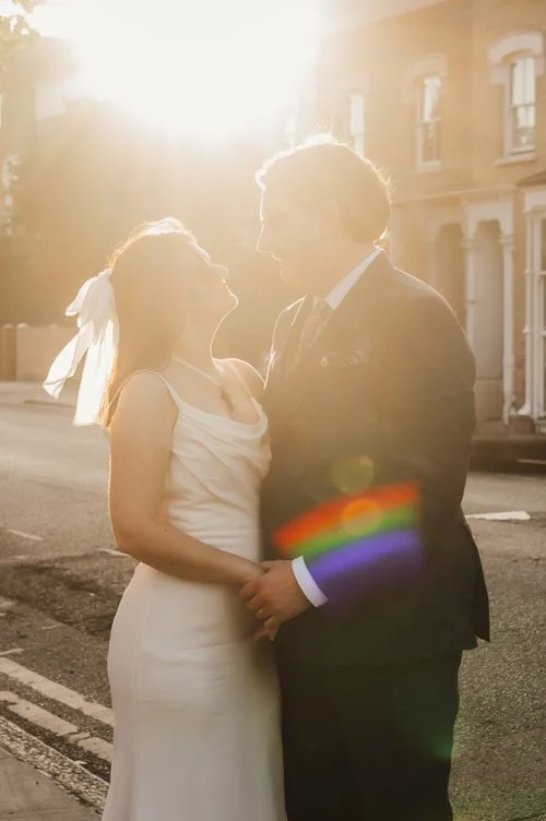 A newly married couple stand together on a quiet London street at golden hour, holding hands as warm sunlight pours over them, creating a soft, romantic glow and rainbow lens flare. The bride’s elegant, simple gown and hair bow catch the light beauti