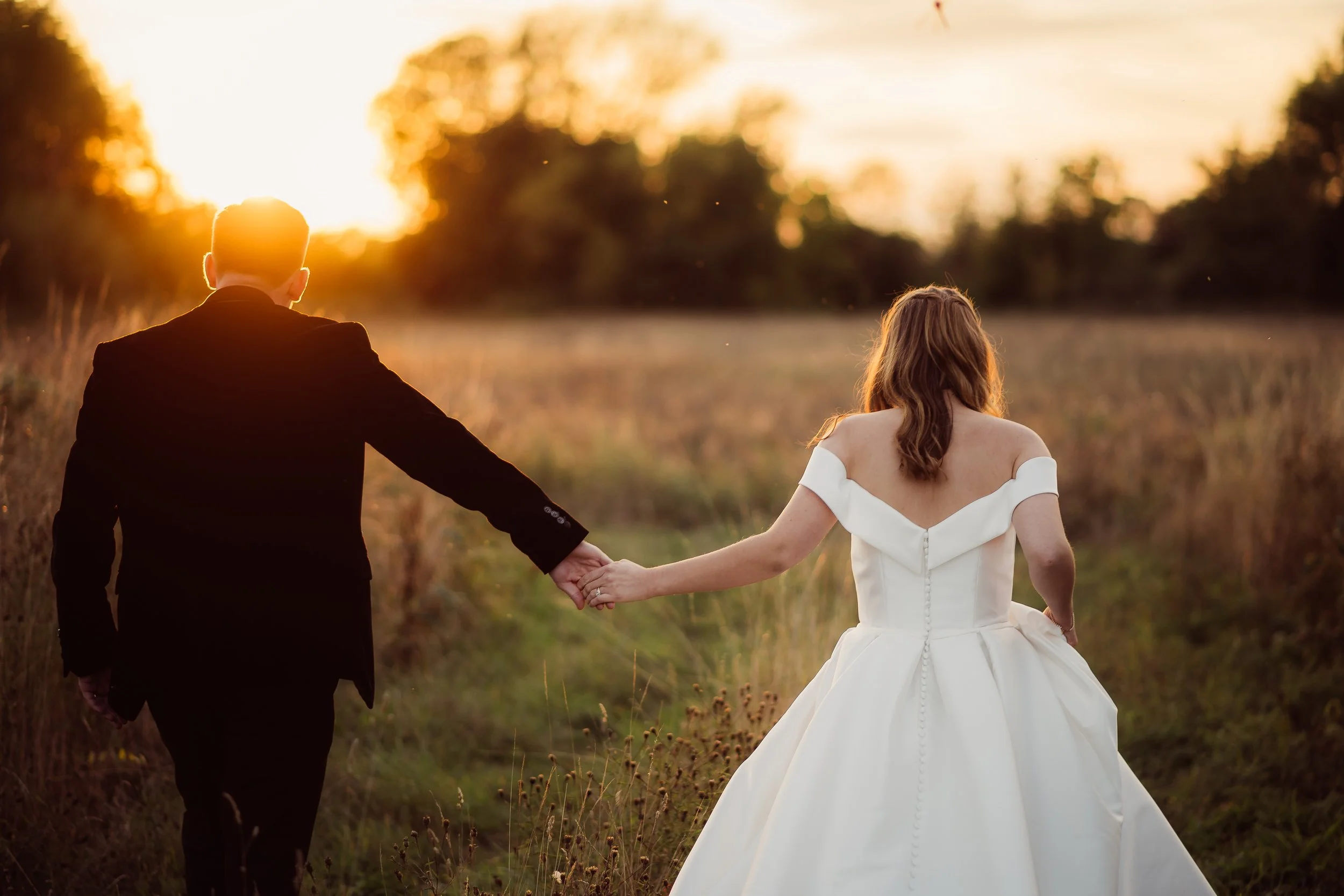 A bride and groom walk hand in hand through a golden field at sunset in this romantic portrait by Gemma Gaskins Photography. The warm backlight, the movement of the bride’s elegant off the shoulder gown and the peaceful countryside setting create a t