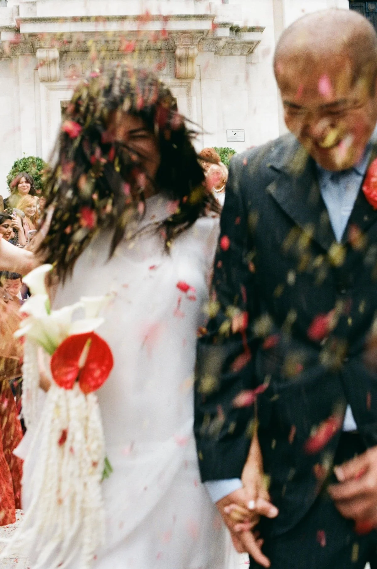 A blurred photo of a bride and groom being scattered with confetti. The bride is holding a simple bouquet with a shiny, red anthurium by Kitty Willow Flower Studio. The images was taken by Emily Krouse.