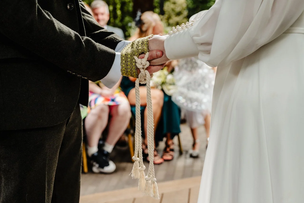 The couple’s hands are gently bound with an ornate cord that drapes between them while sunlight catches the gold detail on their cuffs and the soft fabric of the bride’s sleeves creating an intimate close up of their handfasting that feels tender sym
