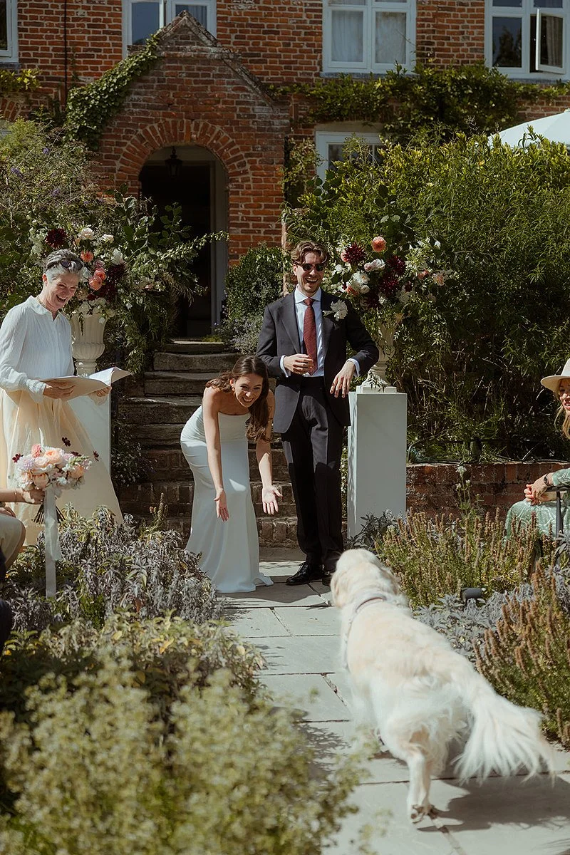Arabella stands to the left holding an open book as the couple lean forward on the garden path, smiling and gesturing toward a dog approaching them. The scene is set outside a brick building framed by greenery and floral arrangements, capturing a liv