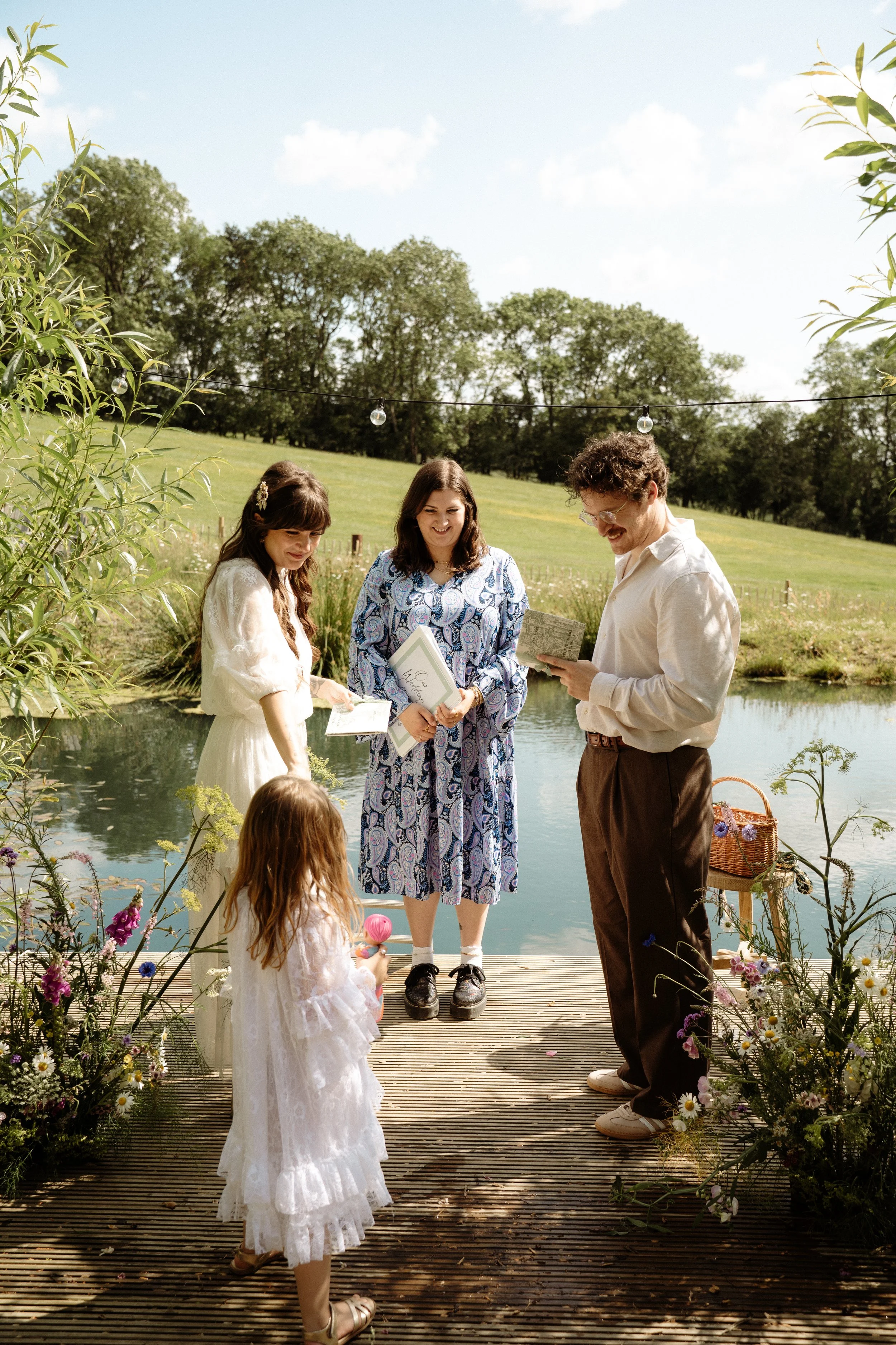 A relaxed and intimate outdoor ceremony beside a serene pond. Lauren The Spiritual Celebrant stands in the centre, holding a ceremony book, while a couple faces each other with joyful expressions, exchanging what appear to be vows or readings. A youn