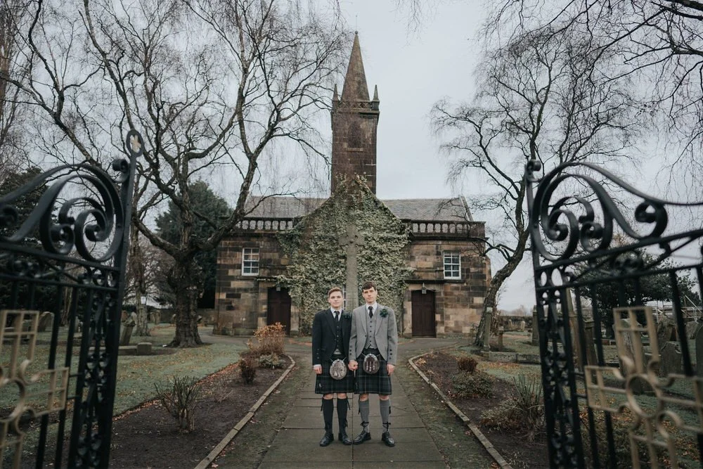 Two grooms wearing traditional Scottish kilts stand hand in hand outside a historic church, framed by wrought iron gates in a striking Glasgow wedding portrait.