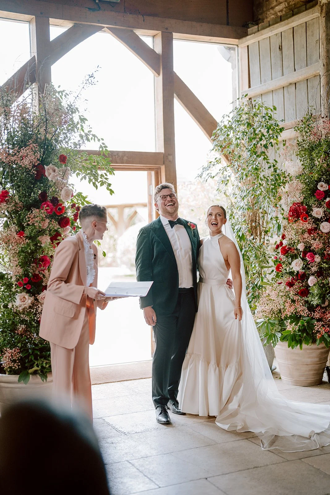 A wedding ceremony in a rustic UK barn setting, with a smiling couple sharing a laugh with modern wedding celebrant Chloe Green. The bride looks radiant in a chic halter-neck gown, while the groom wears a stylish dark green velvet suit. Surrounded by