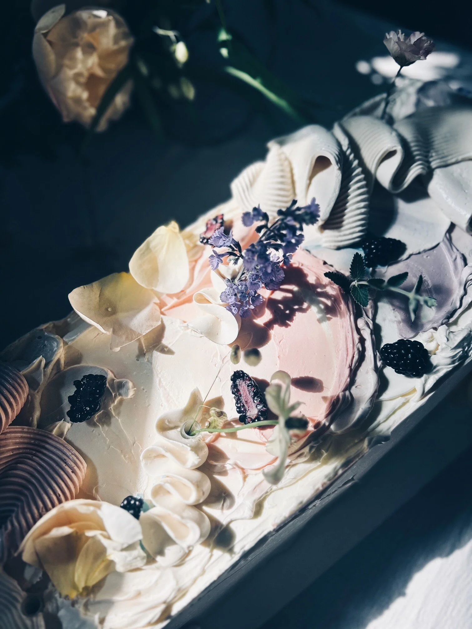 A close up of a long, tray cake by Sarah and Cake. The sun is hining on the cake and there are flowers and fresh fruit pieces.