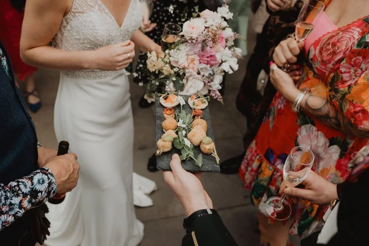 Wedding guests enjoying elegant canapés served by Regis Banqueting at a stylish reception. The image captures a beautifully dressed bride with a bouquet of pastel flowers as gourmet bite-sized appetisers are offered on a slate tray, showcasing the ca