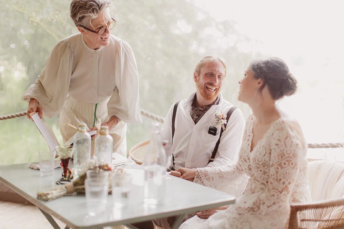 Three people sit and stand around a table in a bright, softly lit setting. One person stands to the left holding papers, while the two seated people face each other, smiling. Glass bottles, drinking glasses, and small decorative items are arranged on