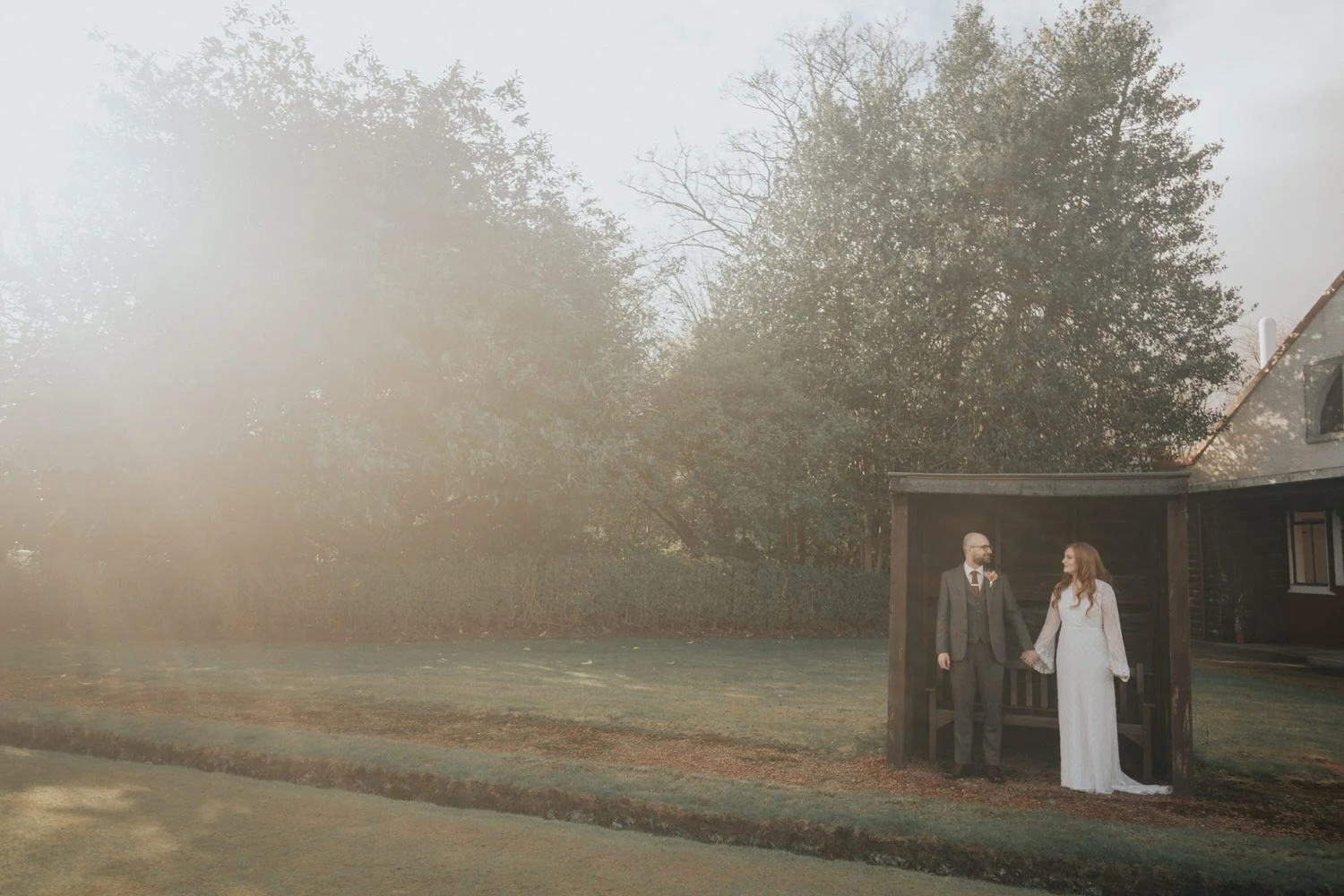 A newlywed couple stand hand in hand beneath a rustic wooden shelter, framed by soft morning haze and winter trees, captured in a relaxed, cinematic moment by Good Luck Wolf wedding photography in Scotland.