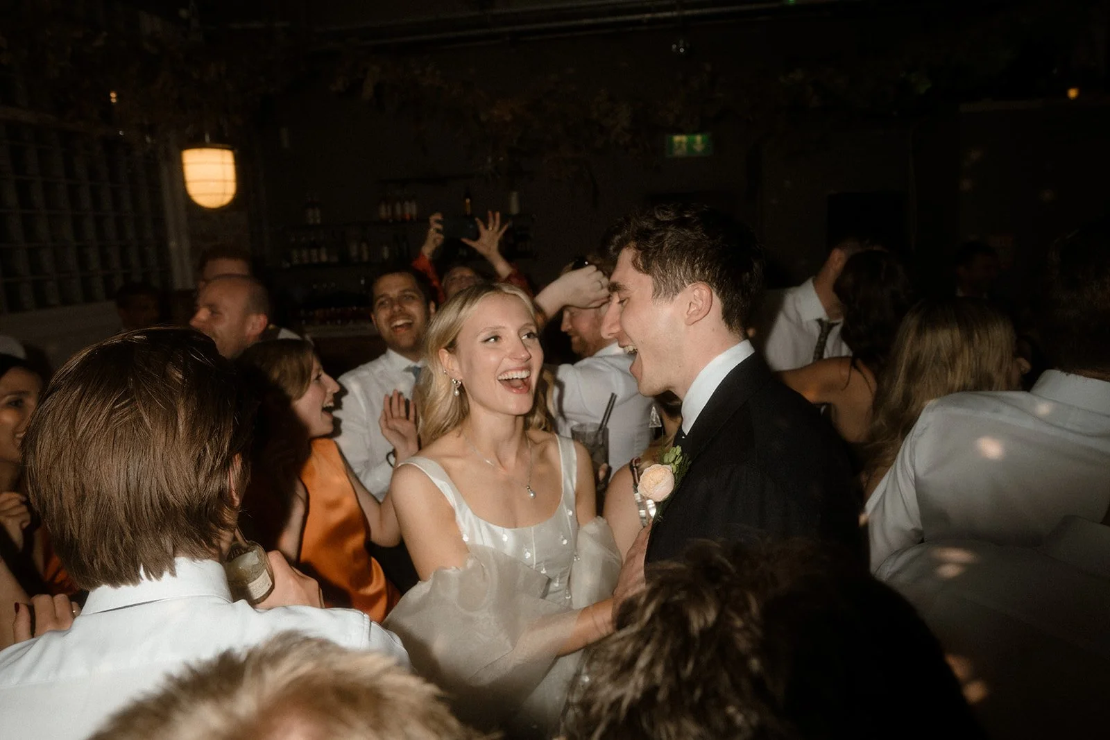 A bride and groom are surrounded by cheering wedding guests on a crowded dance floor as they smile and laugh together during the reception. The bride wears a white dress with sheer sleeves and the groom wears a dark suit with a boutonniere while peop