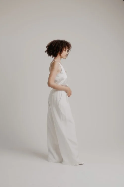 A woman stands in profile wearing a white sleeveless bridal jumpsuit with wide-leg trousers against a soft neutral studio backdrop.