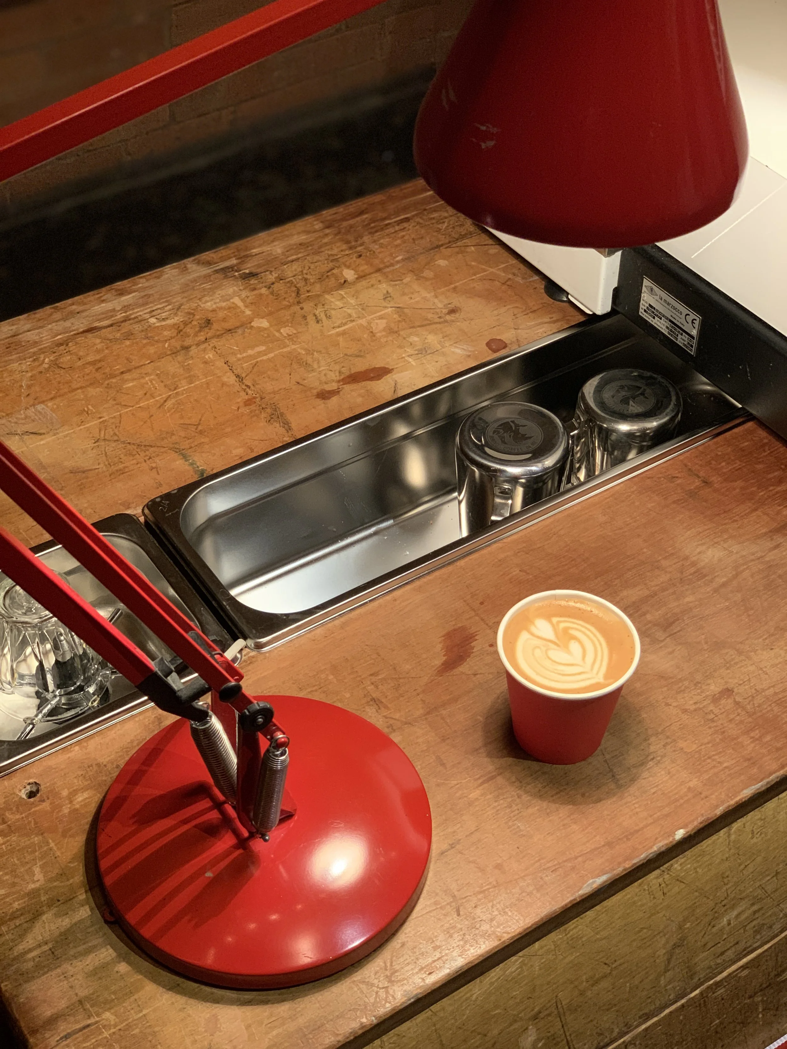 A cosy Red Panda Coffee moment featuring a perfectly poured latte in their trademark red cup, sitting on a well-loved wooden counter illuminated by a bold red lamp with the espresso setup tucked neatly beneath it creating a warm handcrafted atmospher