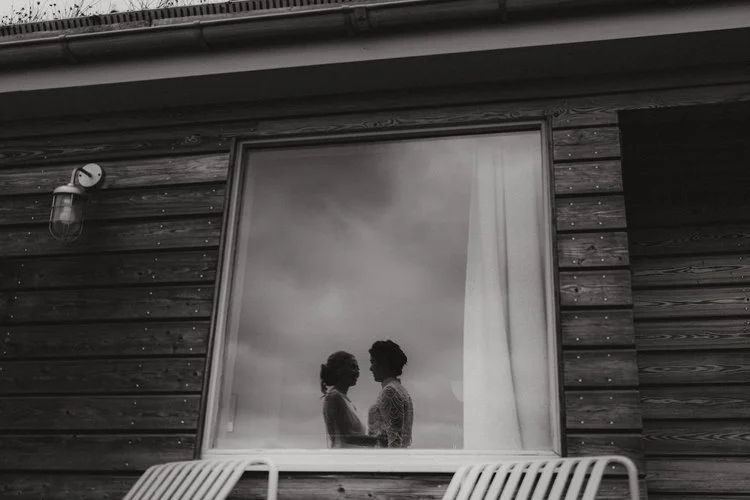 A quietly powerful moment captured by F.D Young Photography: two brides standing close inside a window-framed scene, their silhouettes softened by moody light and drifting reflections. The black-and-white tones draw attention to their connection, tur