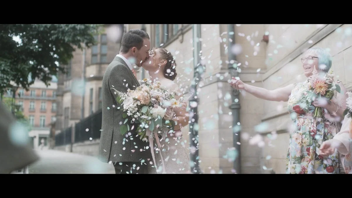 Soft pastel confetti fills the air as the couple share a tender kiss outside a grand stone building while the bride’s bouquet of muted blooms rests between them and joyful guests shower them in colour creating a dreamy cinematic moment.