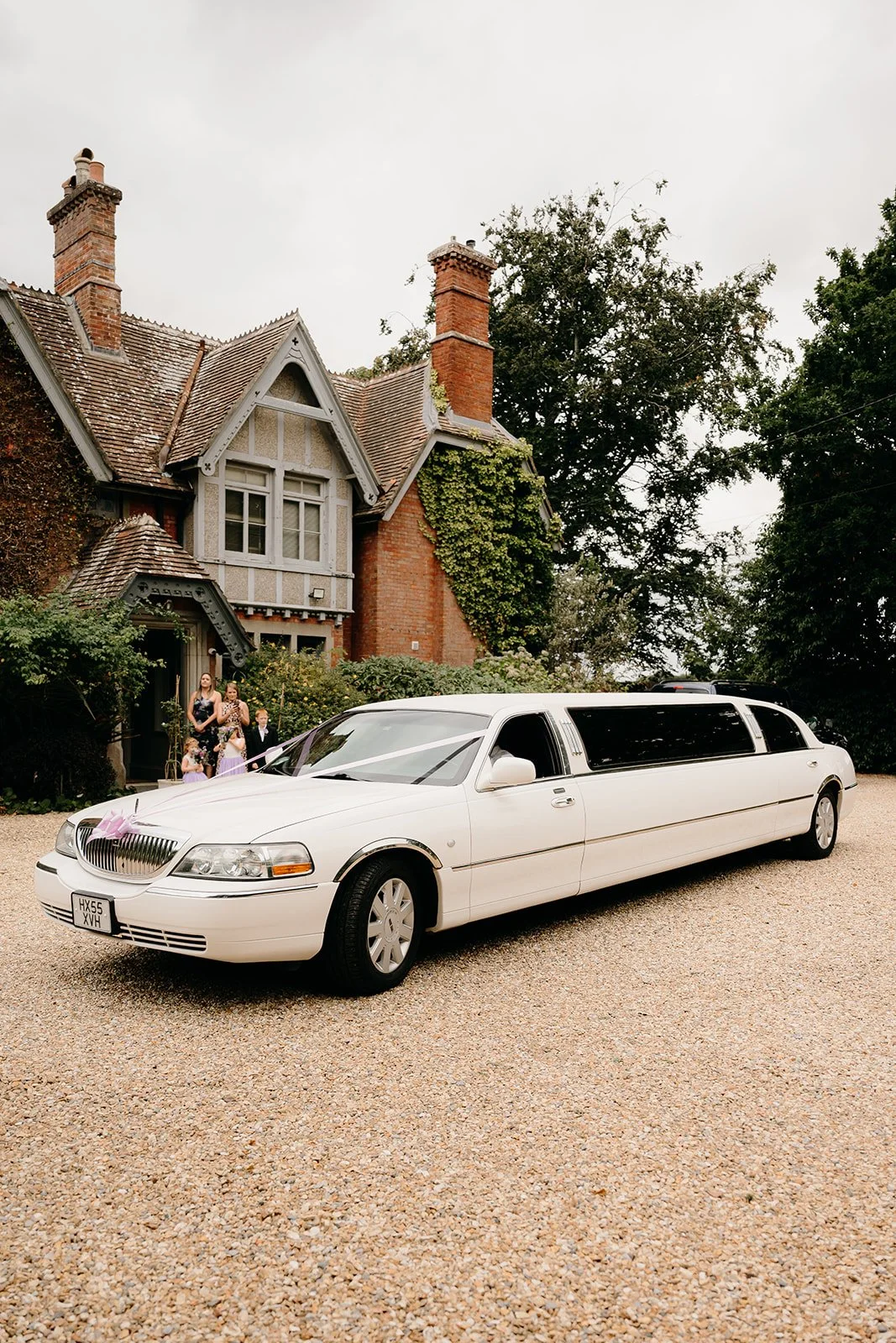 A white limousine is parked outside The Old Vicarage in Hampshire. There is a lilac bow on the front and some wedding guests waiting patiently.