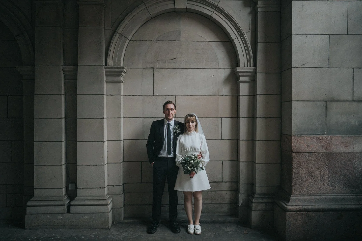 A relaxed Glasgow city wedding portrait of a couple standing beneath a grand stone archway, with the bride in a short white dress and veil holding a bouquet and the groom in a dark suit, captured in a moody, documentary style by Good Luck Wolf weddin
