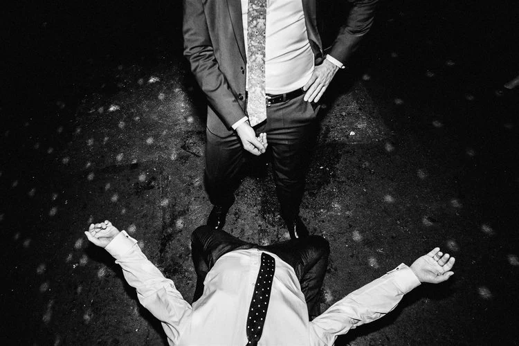 A sharply lit black and white photo that captures a chaotic and humorous dance floor moment from an overhead angle. One person lies flat on their back on the ground with arms flung wide, shirt rumpled, and tie falling off-center. Standing above them 