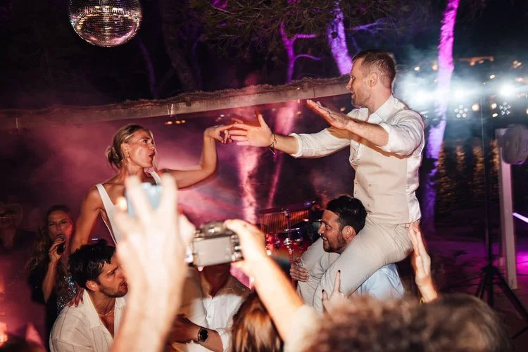 Guests lifting Mr and Mrs L onto shoulders during an outdoor wedding party, surrounded by colourful lighting, smoke effects, and disco balls. The energetic scene captures the excitement of a packed dance floor, perfect for couples wanting unforgettab