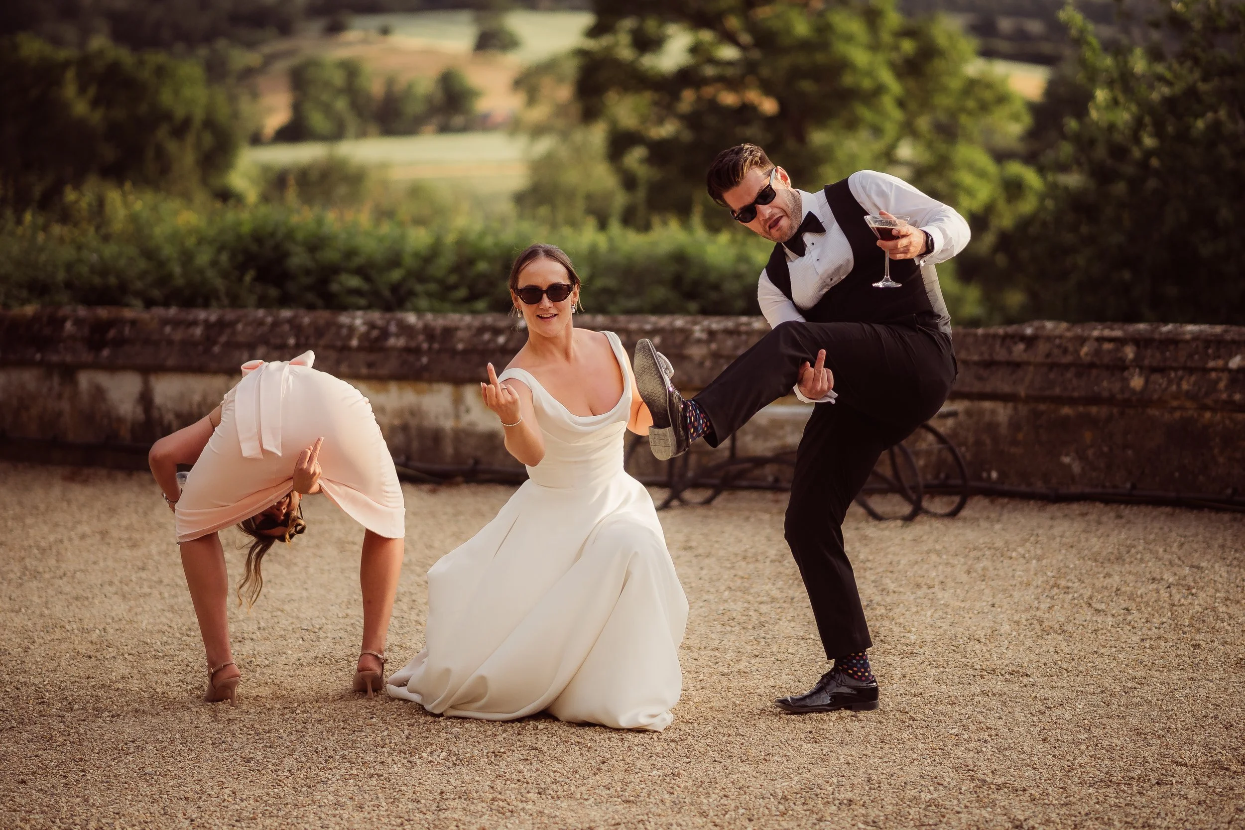 A playful candid wedding photo showing three adults having fun outdoors during golden hour. The bride in a modern white wedding dress and sunglasses kneels on a gravel courtyard while making a cheeky hand gesture. A bridesmaid in a pale pink dress be