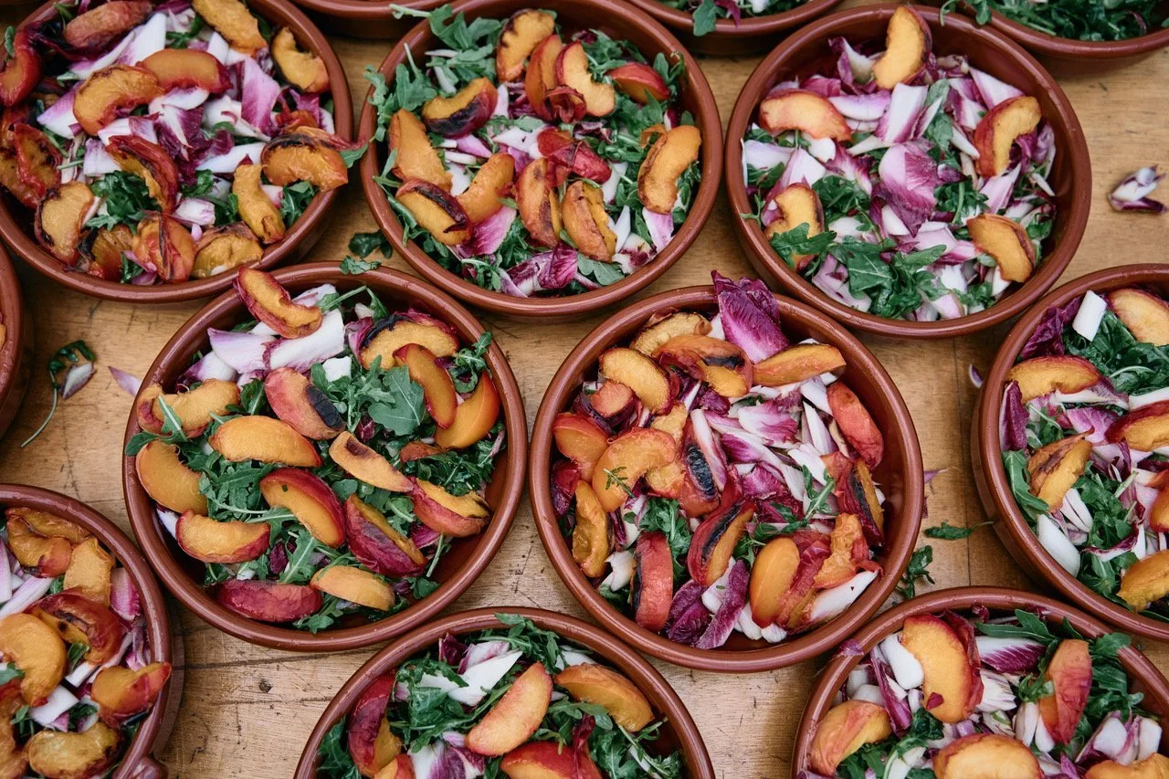 A colourful selection of freshly prepared wedding salads by Tasting Plates Wedding Caterers, featuring vibrant bowls filled with rocket, radicchio, and juicy roasted peaches. Each dish is beautifully arranged in rustic terracotta bowls, showcasing Ta