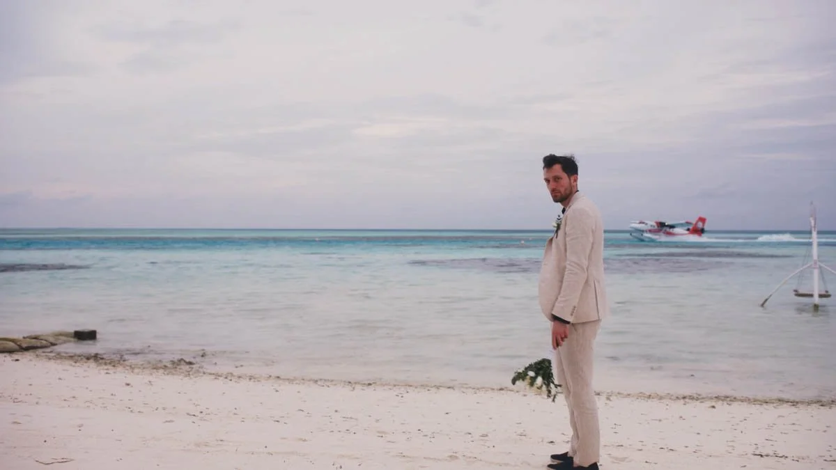 A groom stands alone on a pale shoreline with the soft pastel sky mirrored in the calm turquoise water behind him while he holds a loose bouquet at his side and the distant seaplane adds a cinematic sense of place that feels both romantic and quietly