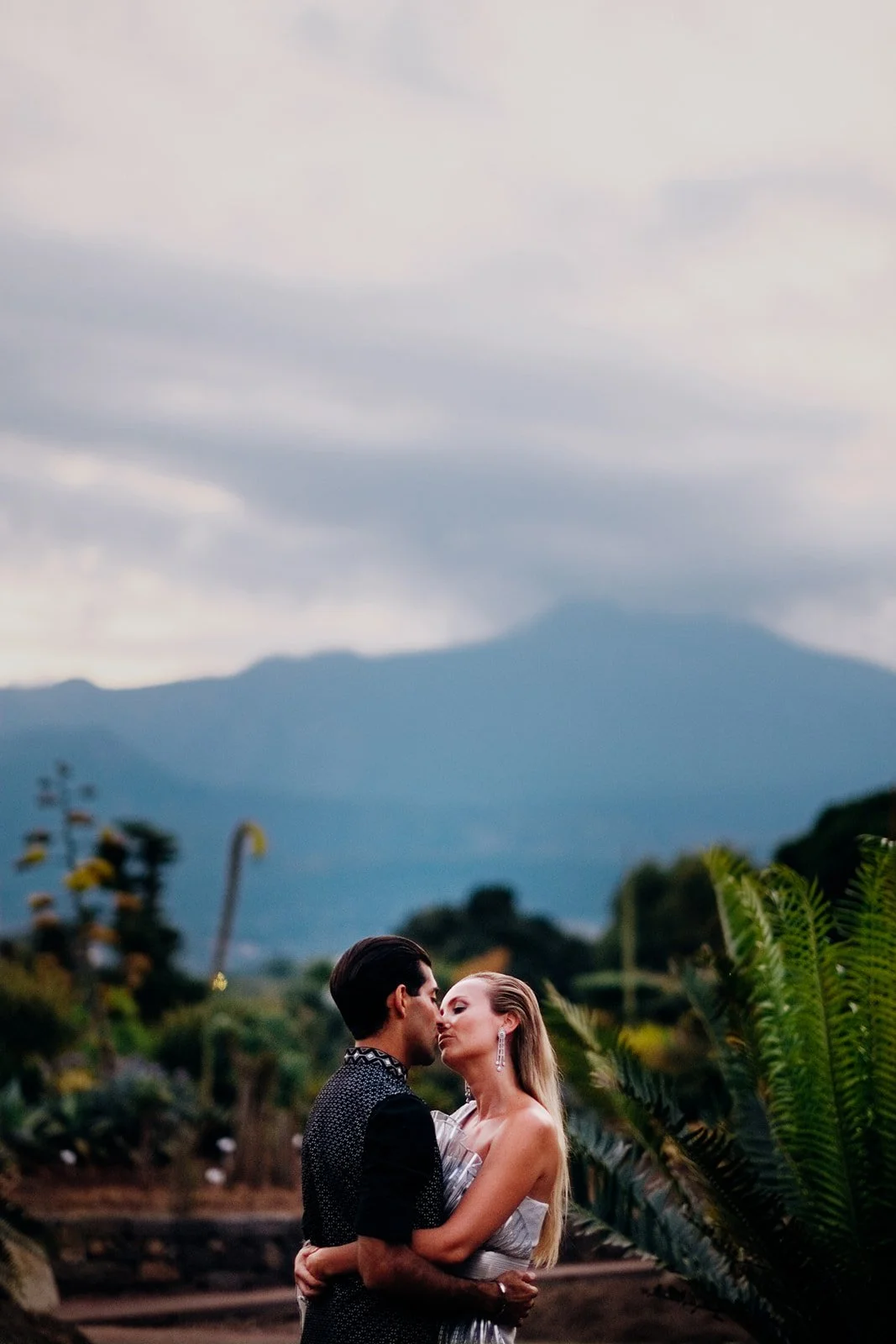 A couple stands closely embracing in an outdoor landscape at dusk, with mountains and a cloudy sky forming a soft, muted backdrop. The person on the left wears a dark patterned outfit while the person on the right, dressed in a light metallic-looking
