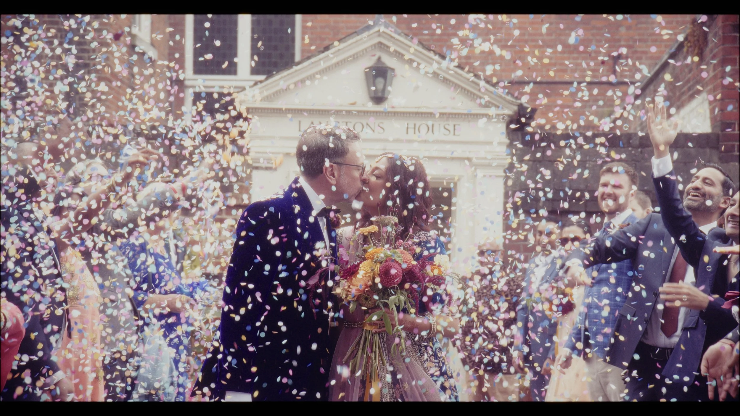 A joyful confetti-filled moment captured by Heart Shaped Video, featuring a newly married couple sharing a kiss outside Langtons House surrounded by cheering guests. The image bursts with colour and emotion - confetti swirling in the air, a bright br
