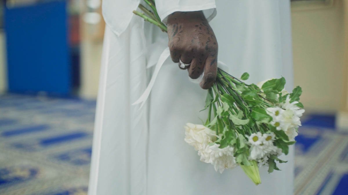 In this quiet atmospheric moment a hand adorned with intricate henna rests gently at the side holding a small bouquet of white flowers with soft petals and fresh green stems while the flowing white fabric of the outfit drapes gracefully toward a patt