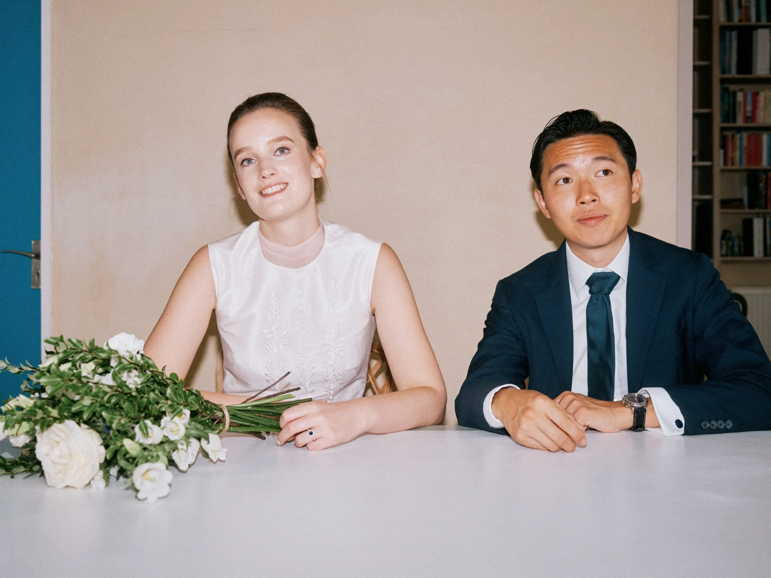 A stylish and modern wedding portrait captured by Wayne La Photography, showing the couple seated together at a simple white table in a quiet, softly lit room. The bride’s sleeveless ivory dress and relaxed posture create a sense of ease, while the g