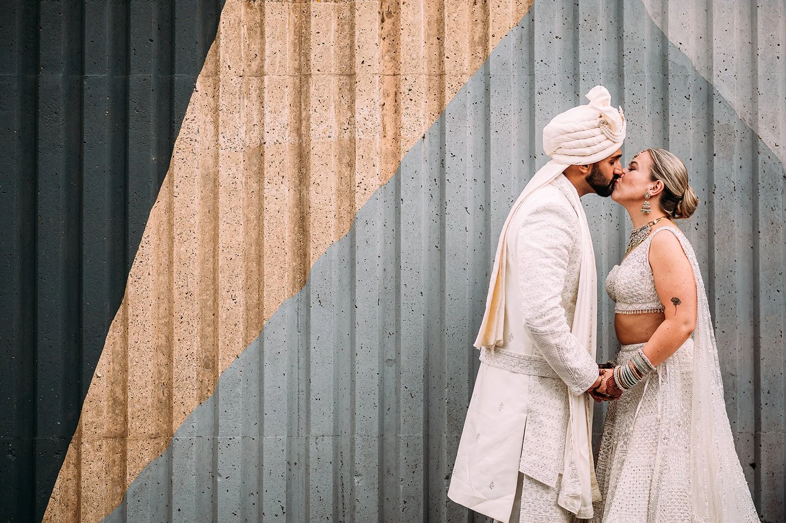 A couple dressed in intricately embroidered ivory wedding attire share a gentle kiss while holding hands in front of a corrugated metal wall painted with bold geometric shapes in muted blue, sand, and grey tones, creating a striking contrast between 