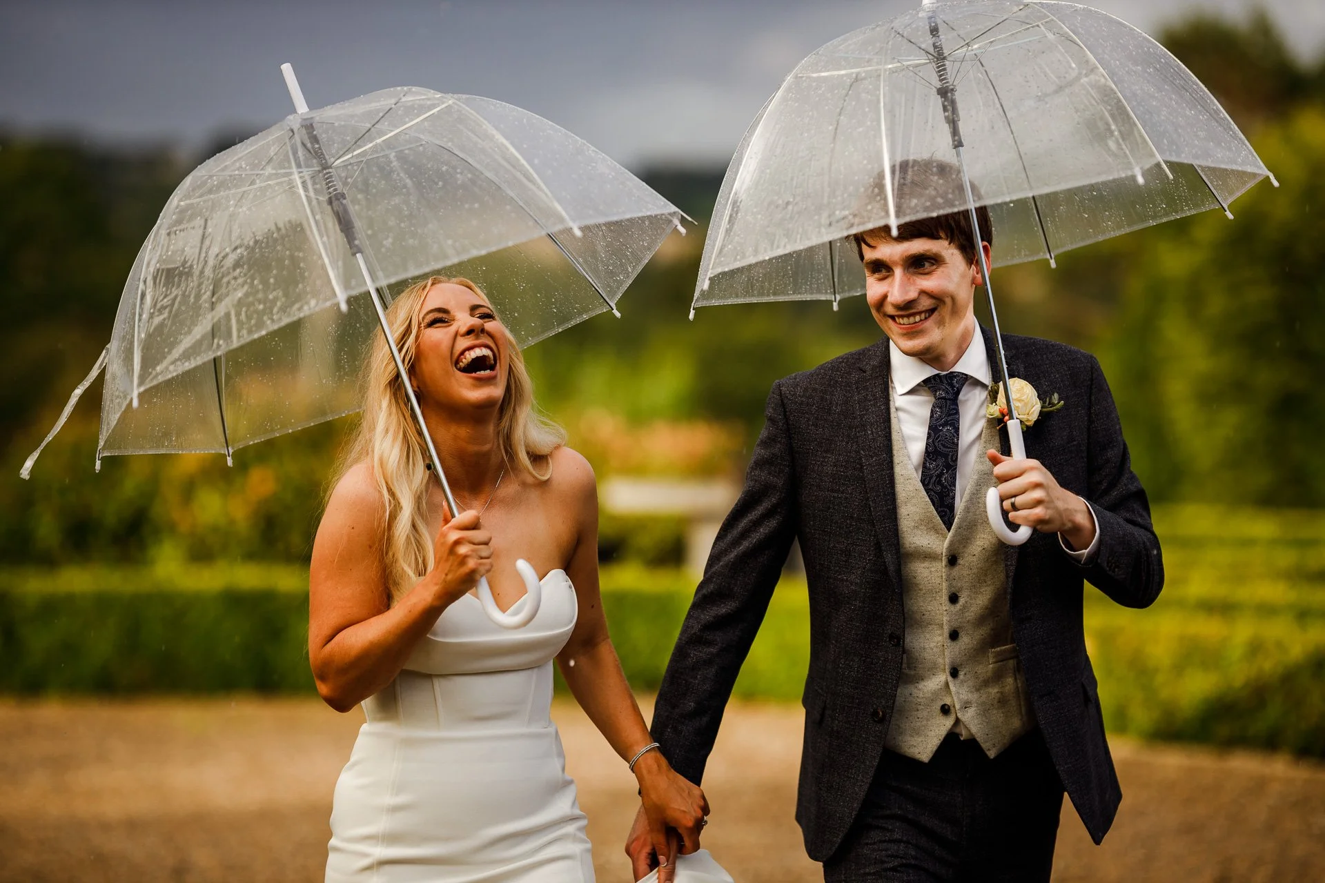 A joyful bride and groom walk hand-in-hand under clear umbrellas during a rainy UK wedding, laughing together as droplets fall around them. Captured by Bluebell Photo Studio, this candid moment showcases natural, documentary-style wedding photography