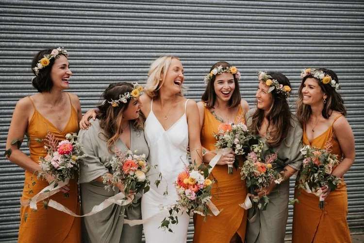 Joyful bridal party holding vibrant seasonal bouquets by Botanique Workshop featuring colourful dahlias, roses and wild garden foliage. With bridesmaids in warm toned dresses and natural floral crowns, this relaxed modern styling highlights Botanique