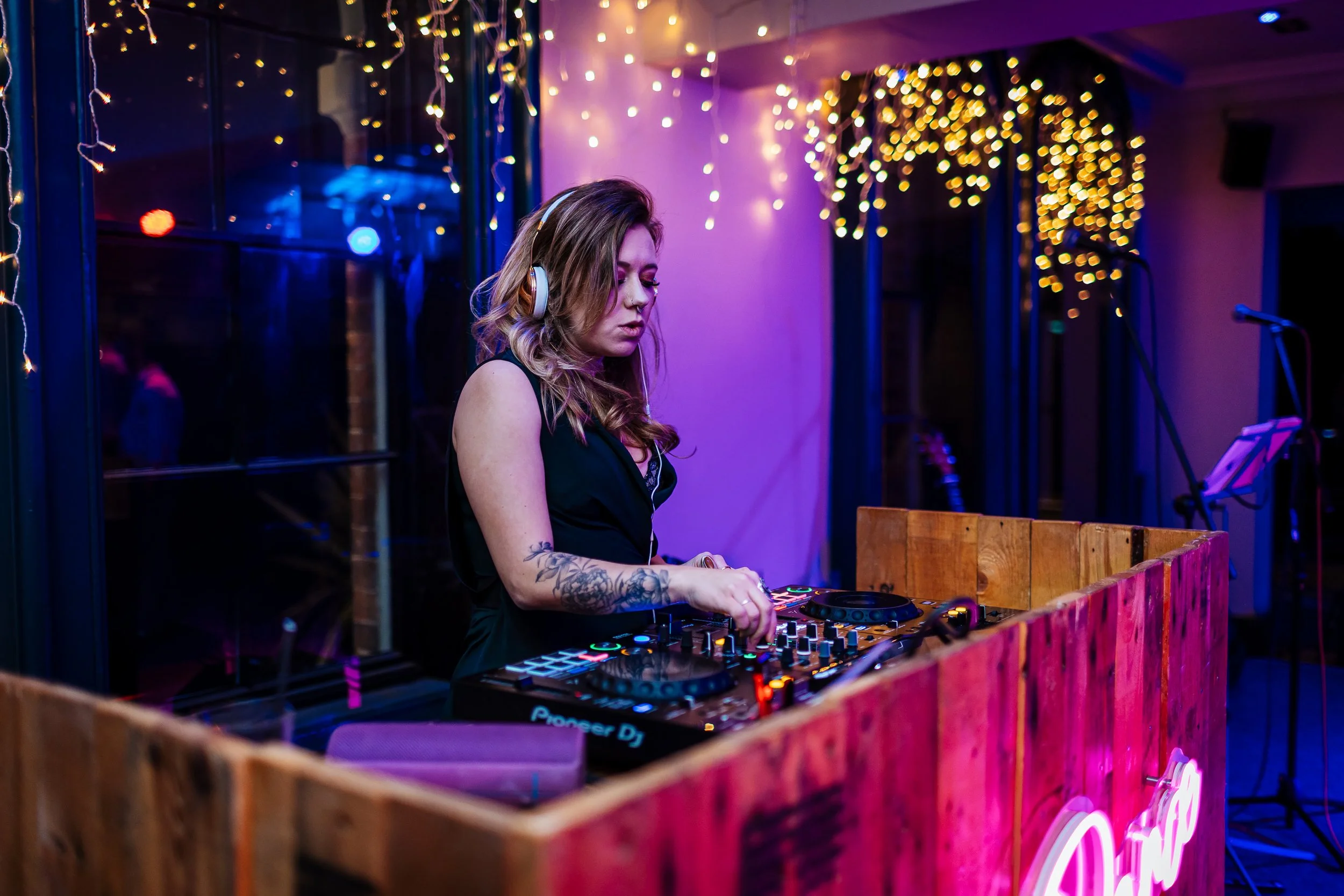 A female wedding DJ from Rave the Date performing at an evening reception, captured by Matt Sim Photography. She stands behind a rustic wooden DJ booth with a glowing neon light, surrounded by twinkling fairy lights. With focused energy and modern st
